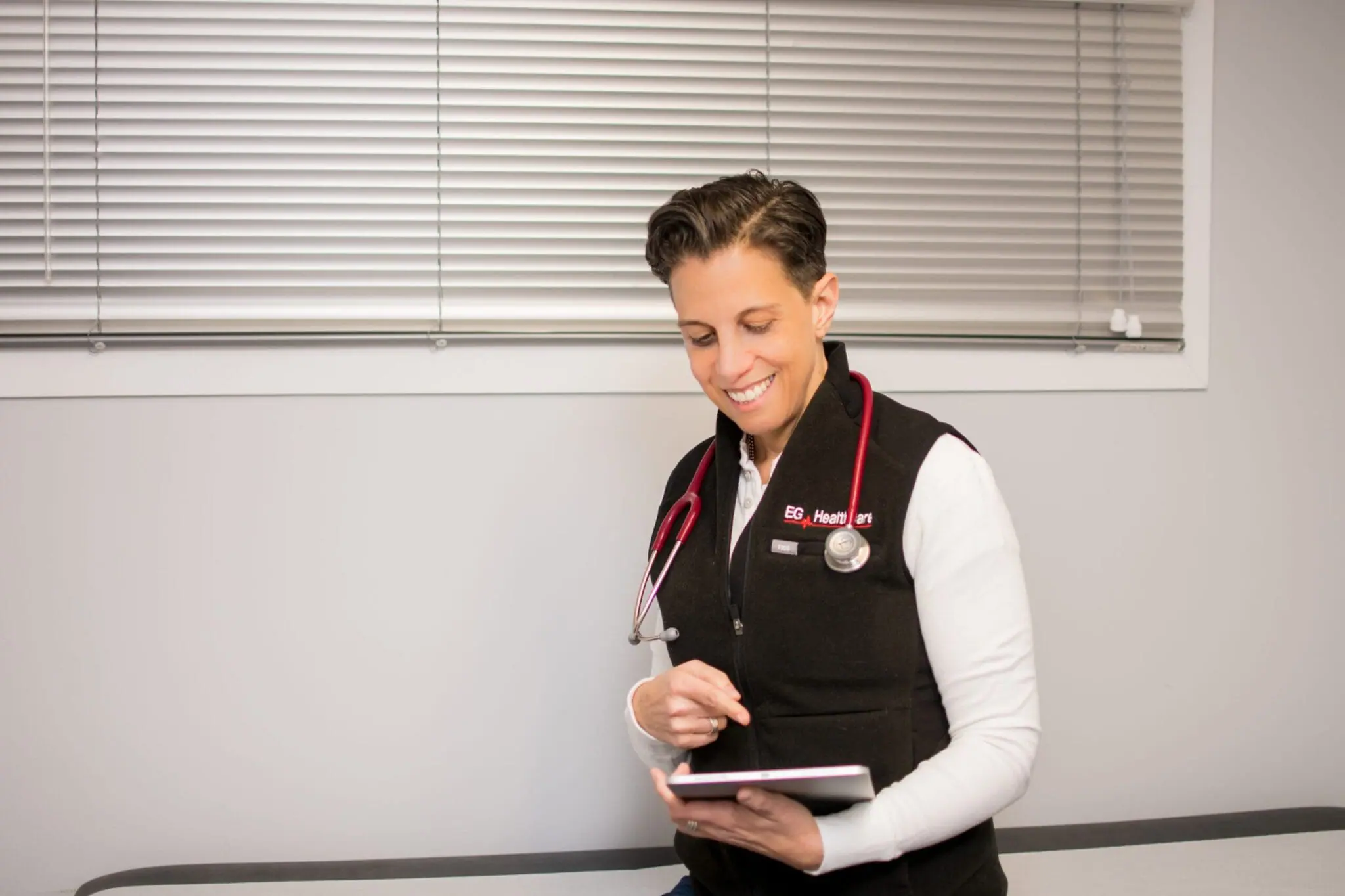 A person wearing a black vest with a stethoscope around their neck stands indoors, smiling and using a tablet, possibly exploring nursing programs.
