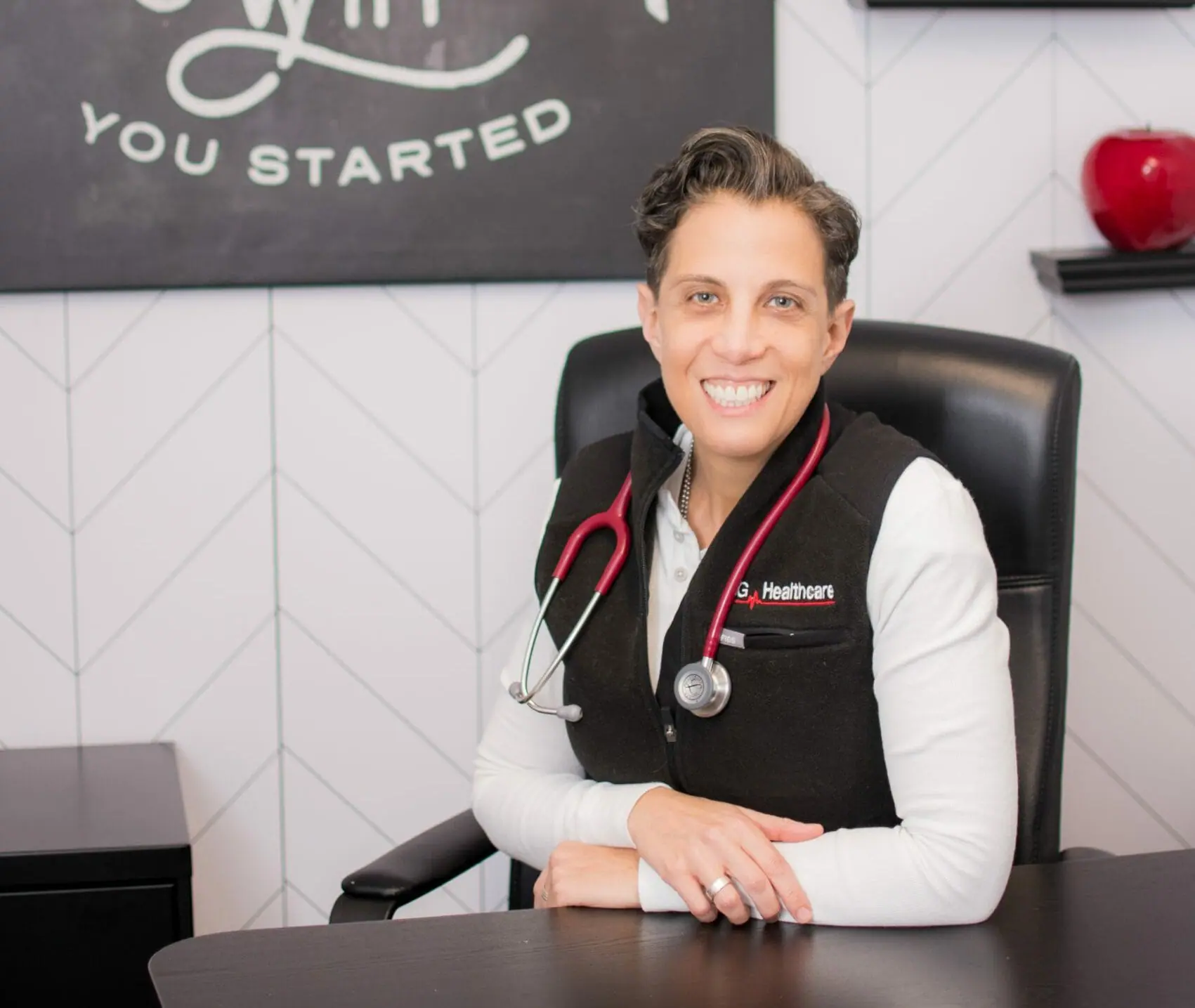 A person seated at a desk with a stethoscope around their neck, smiling. The background features a wall adorned with a motivational message and geometric pattern, reflecting the dedication found in top nursing programs.
